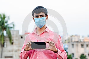 Young Indian man wearing surgical mask and showing smartphone to camera.