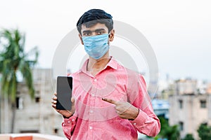Young Indian man wearing surgical mask and showing smartphone to camera.
