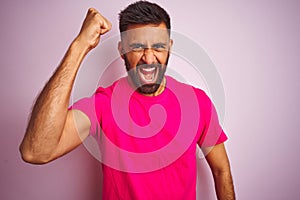 Young indian man wearing t-shirt standing over isolated pink background angry and mad raising fist frustrated and furious while