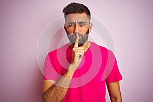 Young indian man wearing t-shirt standing over isolated pink background asking to be quiet with finger on lips