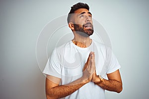 Young indian man wearing t-shirt standing over isolated white background begging and praying with hands together with hope