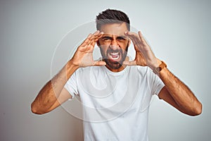 Young indian man wearing t-shirt standing over isolated white background Shouting angry out loud with hands over mouth