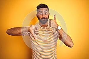 Young indian man wearing t-shirt standing over isolated yellow background Doing thumbs up and down, disagreement and agreement