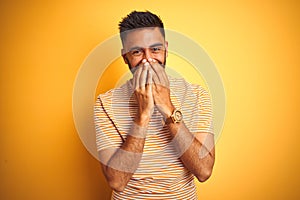 Young indian man wearing t-shirt standing over isolated yellow background laughing and embarrassed giggle covering mouth with