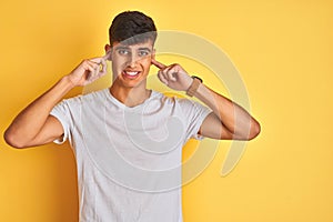 Young indian man wearing white t-shirt standing over isolated yellow background covering ears with fingers with annoyed expression