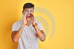 Young indian man wearing white t-shirt standing over isolated yellow background Shouting angry out loud with hands over mouth