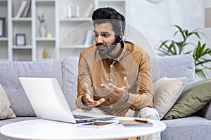 A young Indian man works remotely from home. He sits on the couch in a headset in front of a laptop and chats on a video