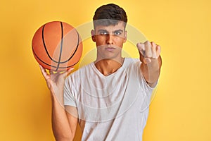 Young indian sportsman holding basketball ball standing over isolated yellow background pointing with finger to the camera and to