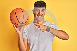 Young indian sportsman holding basketball ball standing over isolated yellow background very happy pointing with hand and finger