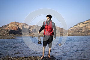 Young indian traveler standing near a lake in the mountains with a lantern, getting ready for camping in the night