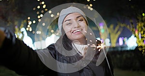 Young Indian Woman making video call holding sparkler standing on lights and garlands background, New Year and Christmas