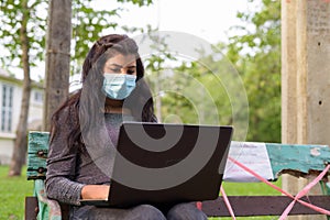 Young Indian woman with mask using laptop while sitting with distance on park bench