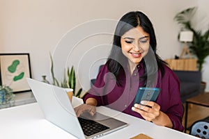 Young indian woman sitting on table at home working on laptop and using phone