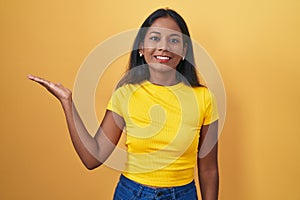 Young indian woman standing over yellow background smiling cheerful presenting and pointing with palm of hand looking at the