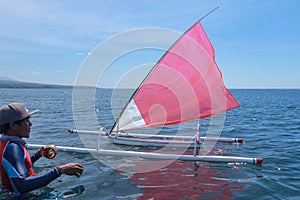 A young Indonesian stands in the sea and prepares a model ship for the competition. Small wooden sailboat with red-white sail