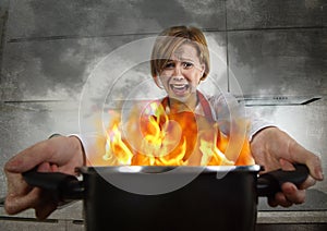 Young inexperienced home cook woman in panic with apron holding pot burning in flames with in panic