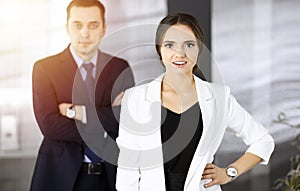 Young intelligent businesswoman is standing straight in a sunny modern office with her colleague at the background