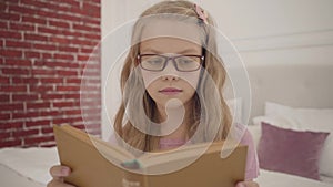 Young intelligent teenage girl in glasses reading the book at home. Smart Caucasian blond schoolgirl studying indoors.