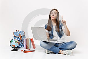 Young interested woman student holding laptop pc computer pointing index finger up sitting near globe, backpack, school