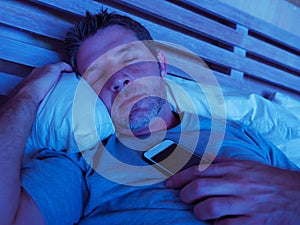 Young internet addict man sleeping on bed holding mobile phone in his hand at night in smartphone and social media network overuse