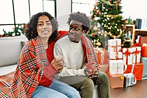 Young interracial couple drinking coffee sitting on the sofa by christmas tree at home