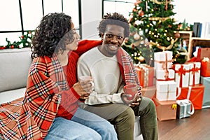 Young interracial couple drinking coffee sitting on the sofa by christmas tree at home