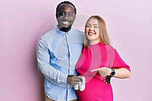 Young interracial couple expecting a baby holding shoes smiling happy pointing with hand and finger