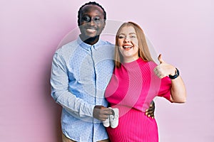 Young interracial couple expecting a baby holding shoes smiling happy and positive, thumb up doing excellent and approval sign