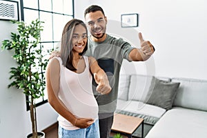 Young interracial couple expecting a baby, touching pregnant belly approving doing positive gesture with hand, thumbs up smiling