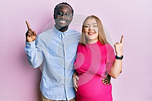 Young interracial couple expecting a baby, touching pregnant belly with a big smile on face, pointing with hand finger to the side