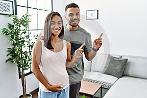 Young interracial couple expecting a baby, touching pregnant belly with a big smile on face, pointing with hand finger to the side