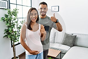 Young interracial couple expecting a baby, touching pregnant belly doing happy thumbs up gesture with hand