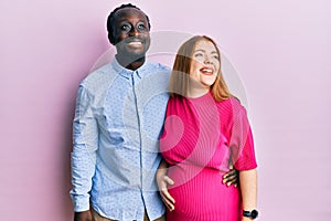 Young interracial couple expecting a baby, touching pregnant belly looking away to side with smile on face, natural expression