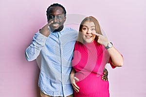 Young interracial couple expecting a baby, touching pregnant belly pointing with hand finger to face and nose, smiling cheerful