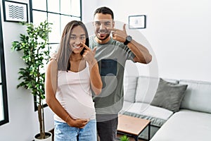 Young interracial couple expecting a baby, touching pregnant belly pointing with hand finger to face and nose, smiling cheerful