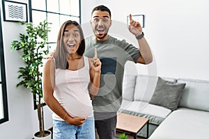 Young interracial couple expecting a baby, touching pregnant belly smiling amazed and surprised and pointing up with fingers and