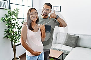 Young interracial couple expecting a baby, touching pregnant belly smiling doing phone gesture with hand and fingers like talking