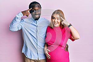 Young interracial couple expecting a baby, touching pregnant belly smiling pointing to head with one finger, great idea or