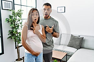 Young interracial couple expecting a baby, touching pregnant belly surprised pointing with finger to the side, open mouth amazed