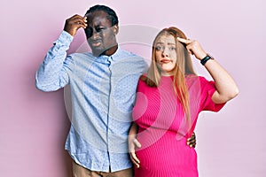 Young interracial couple expecting a baby, touching pregnant belly worried and stressed about a problem with hand on forehead,