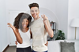Young interracial couple holding keys of new home celebrating victory with happy smile and winner expression with raised hands