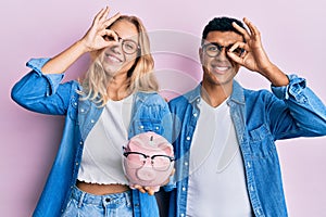 Young interracial couple holding piggy bank with glasses smiling happy doing ok sign with hand on eye looking through fingers