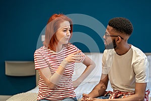Young interracial couple husband and wife sit on bed at home discussing intimacy issues