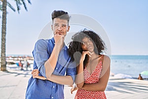 Young interracial couple outdoors on a sunny day with hand on chin thinking about question, pensive expression