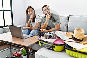 Young interracial couple packing summer clothes in suitcase looking at laptop serious face thinking about question with hand on