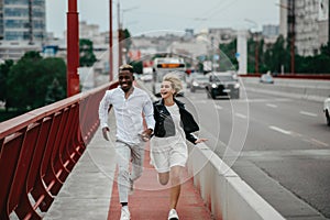 Young interracial couple runs on bridge and laughs cheerfully on the background of the city and the road