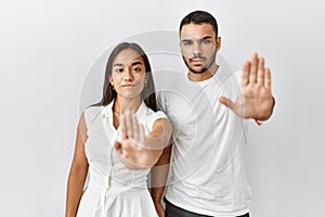 Young interracial couple standing together in love over isolated background doing stop sing with palm of the hand