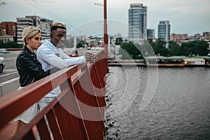 Young interracial couple stands on bridge and hugs against background of river and city