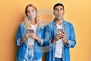 Young interracial couple using smartphone making fish face with mouth and squinting eyes, crazy and comical