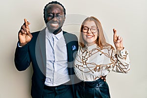 Young interracial couple wearing business and elegant clothes gesturing finger crossed smiling with hope and eyes closed